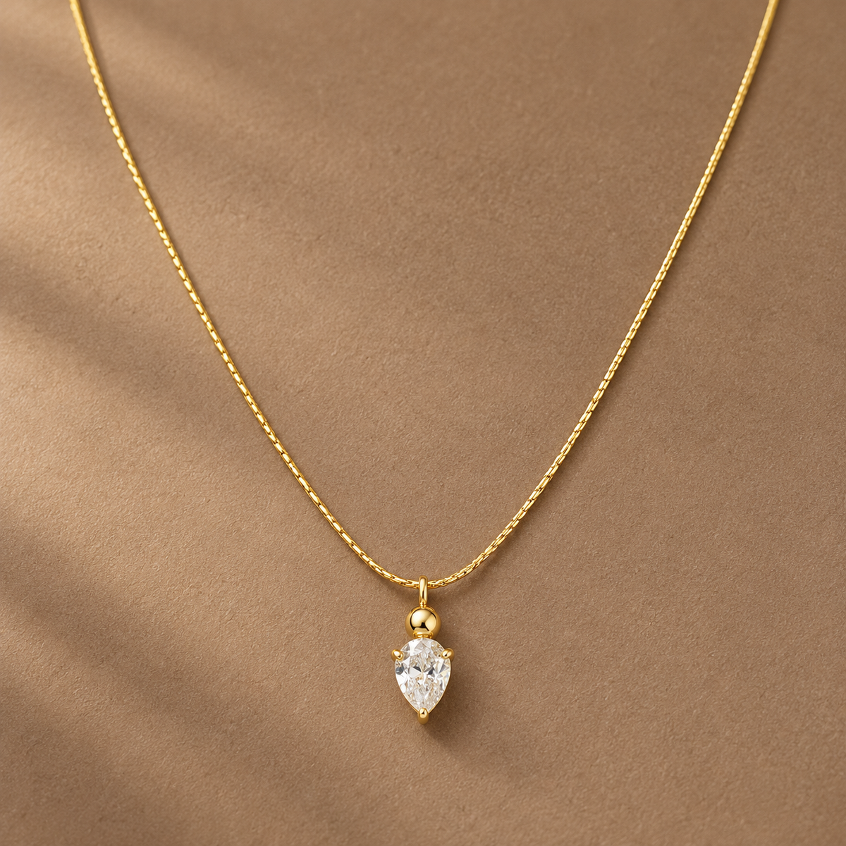 Gold-tone 316L stainless steel necklace with a fine textured chain and a pear-cut clear crystal pendant on a small ball bail, worn at the neckline
