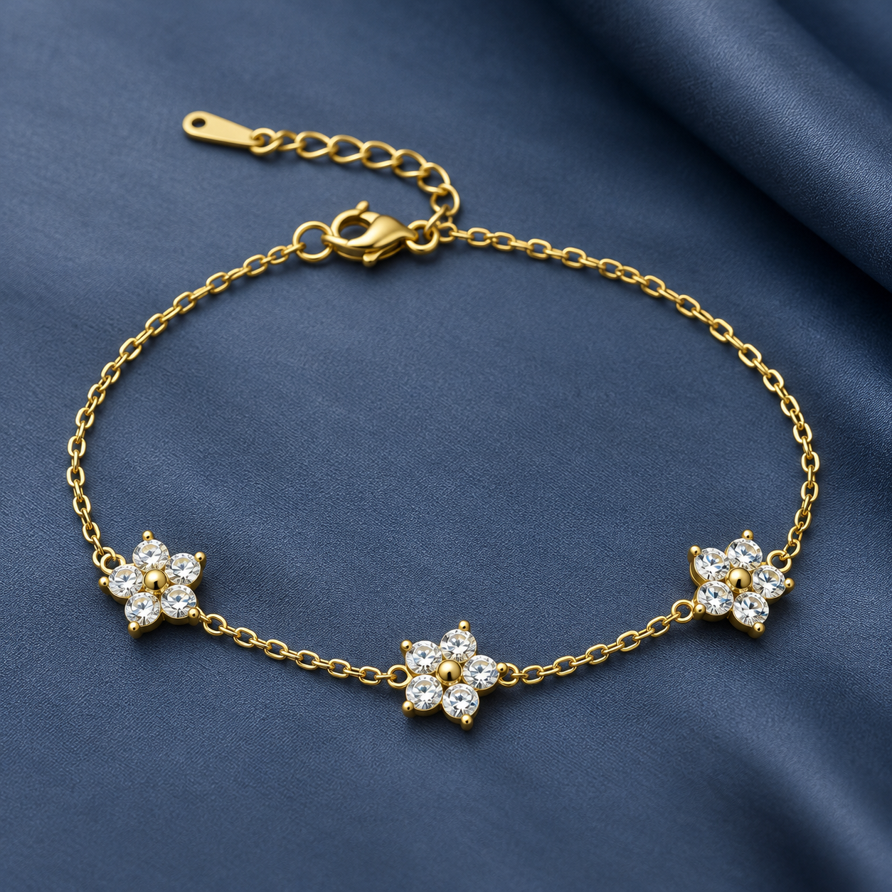 Gold-tone 316L stainless steel chain bracelet with three flower motifs set with clear crystals and a lobster clasp, laid flat on blue fabric