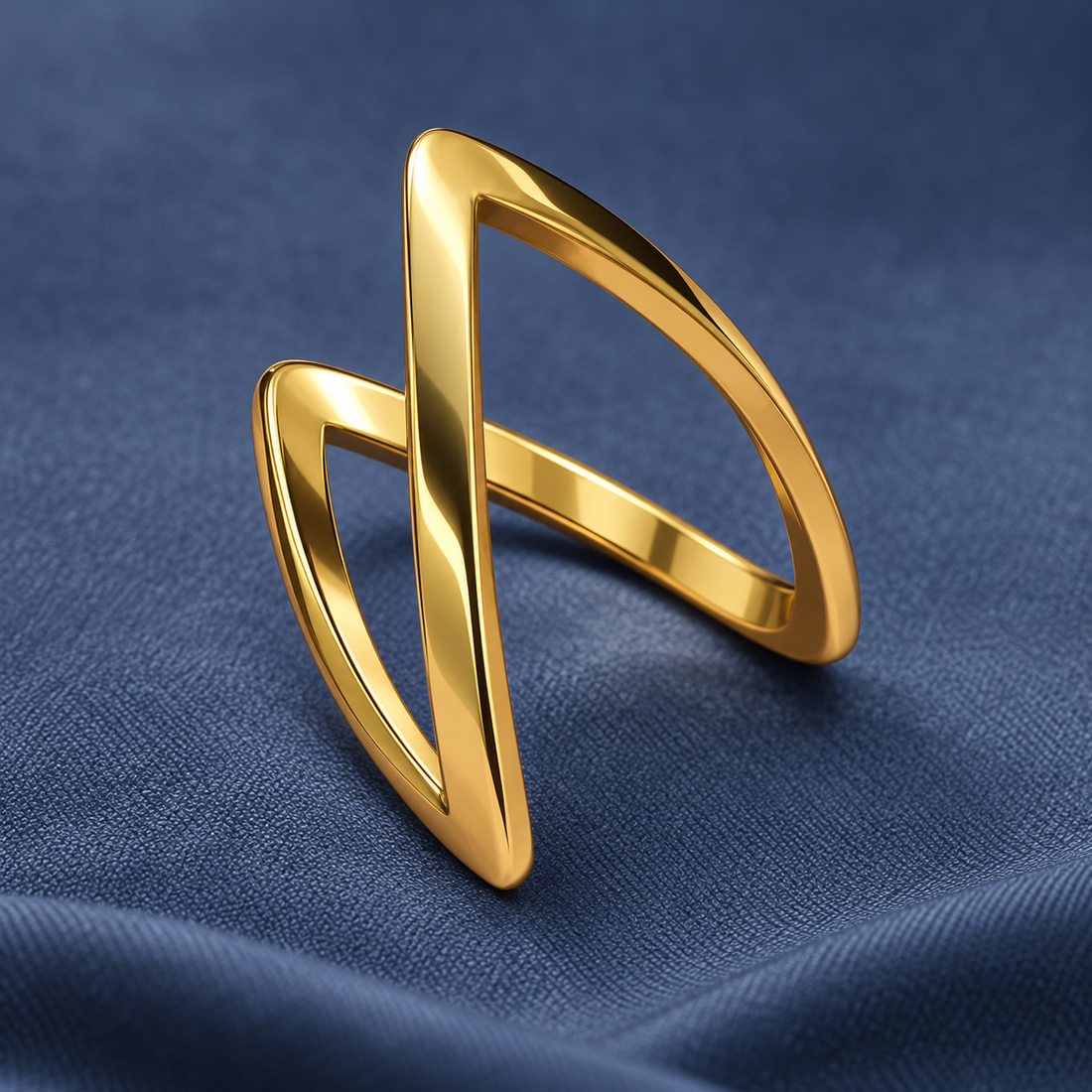 Gold-tone 316L ring with two crossed open bands forming a geometric wrap design, shown on a blue fabric surface