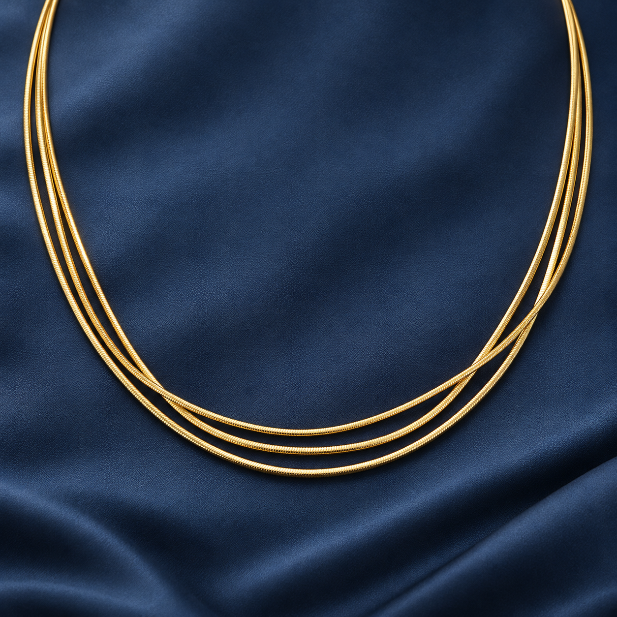 Gold-tone 316L stainless steel necklace of three thin round snake chains laid side by side on a dark blue fabric