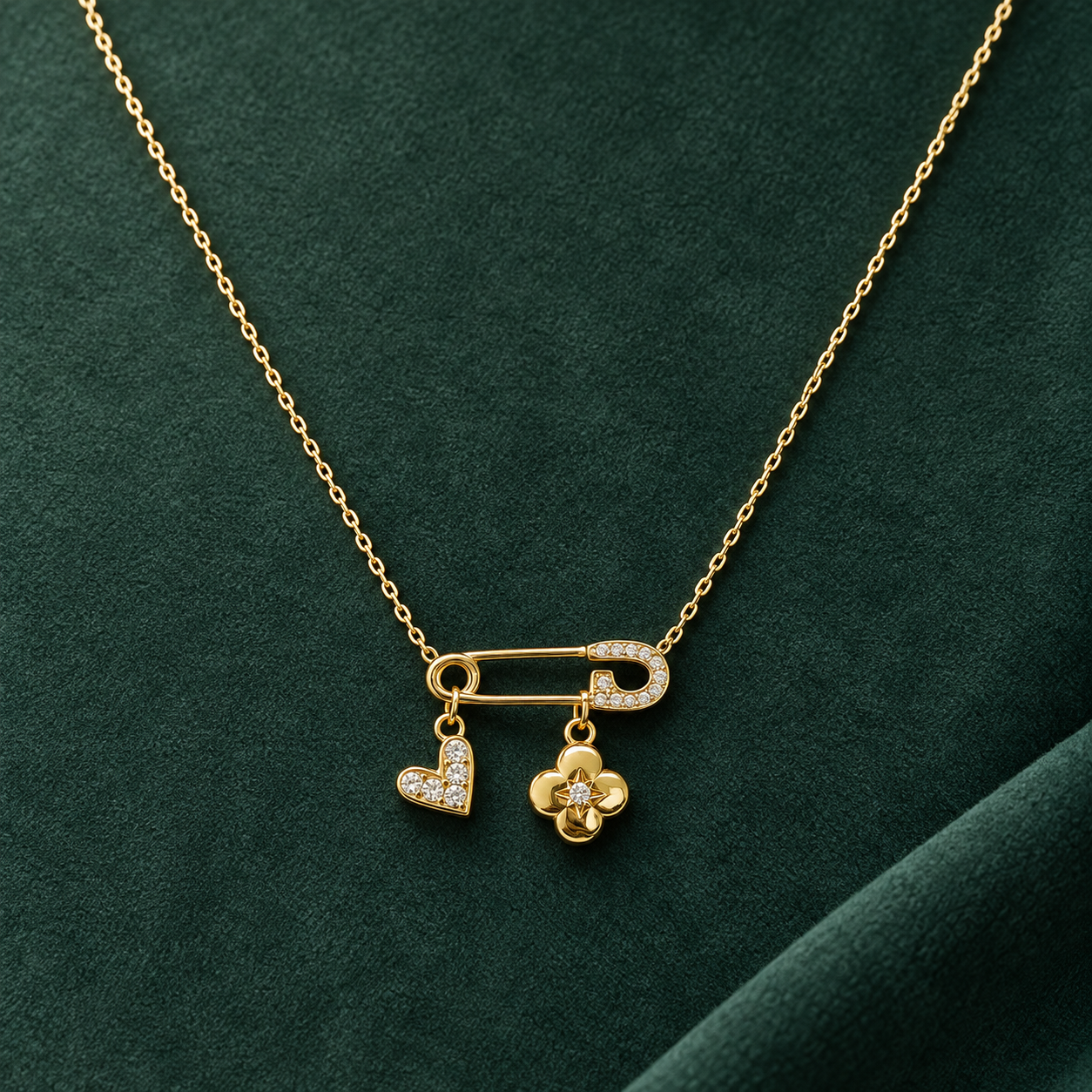 Gold-tone 316L steel necklace with a safety-pin pendant set with crystals, holding a crystal heart and a flower charm, on a green fabric