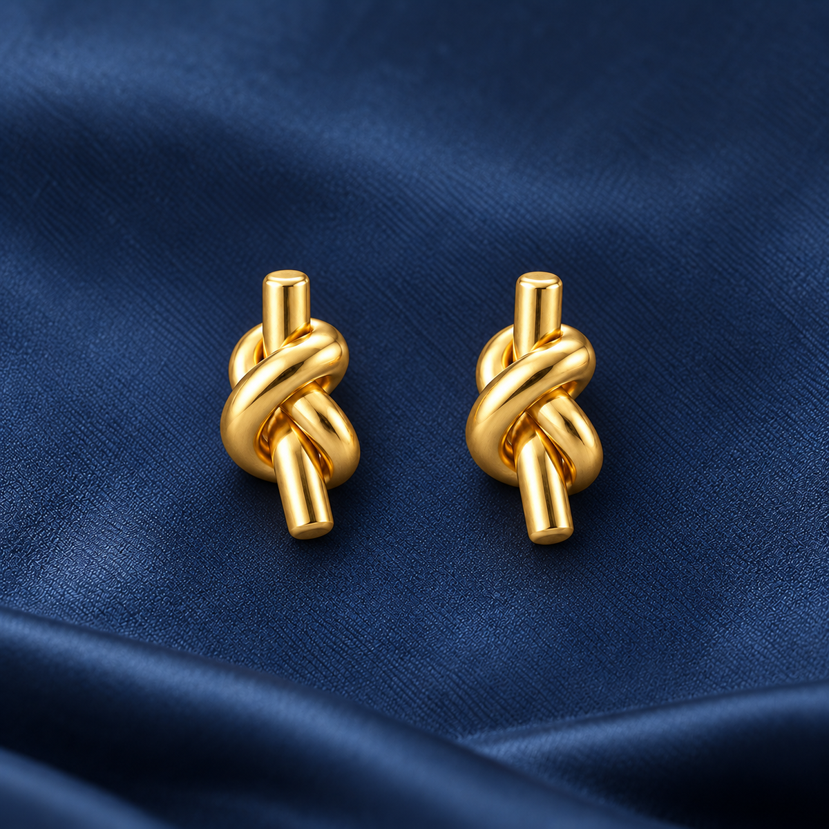 Gold-tone 304 stainless steel pair of earrings with a knotted tube design and straight post backs, shown on a navy fabric