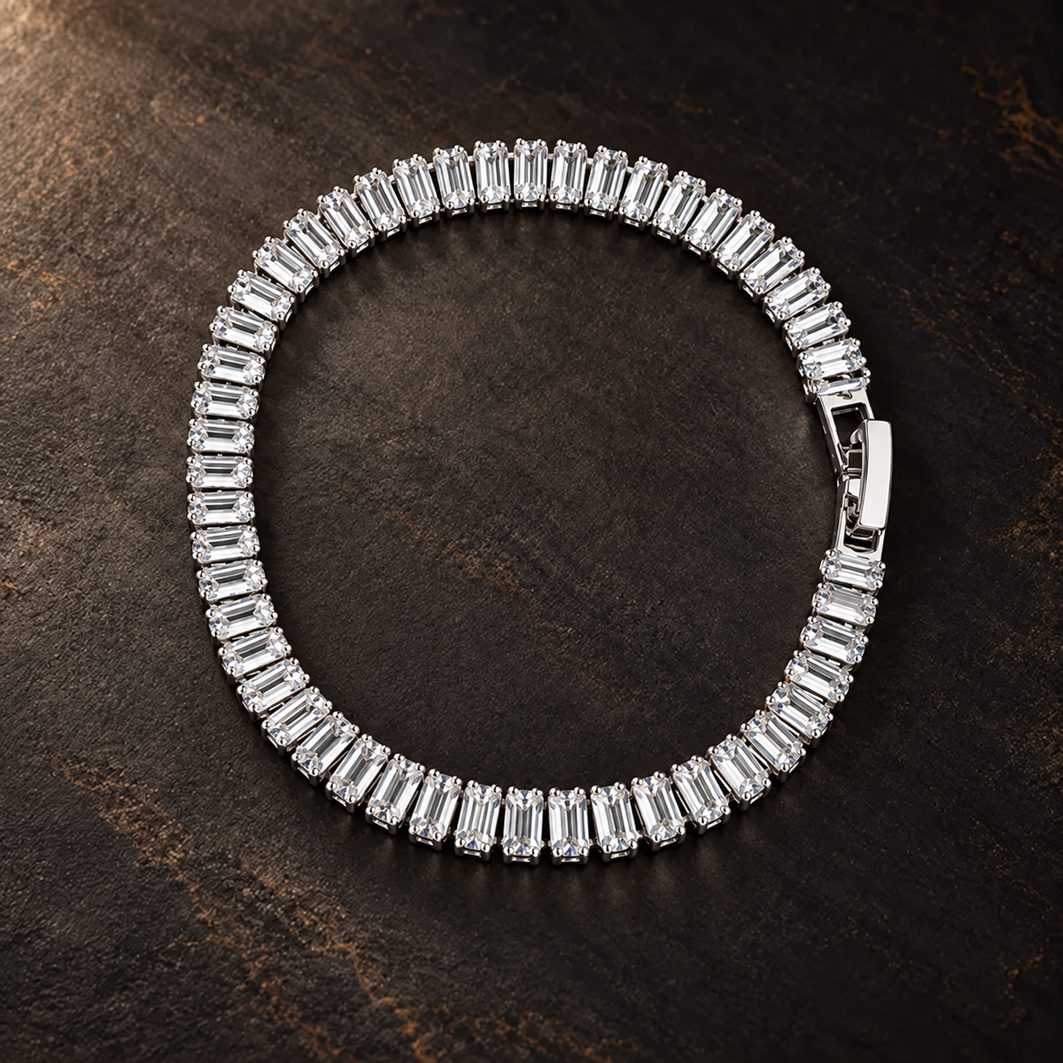 Silver-tone brass tennis bracelet with a row of baguette-cut clear stones in claw settings and a box clasp, laid flat on a dark surface