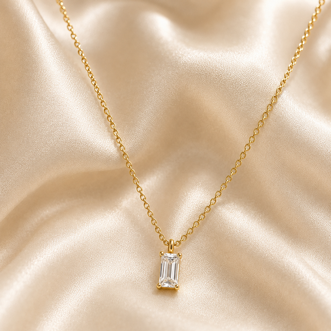A gold chain necklace with an emerald-cut clear gemstone pendant, displayed on a draped champagne satin fabric background.