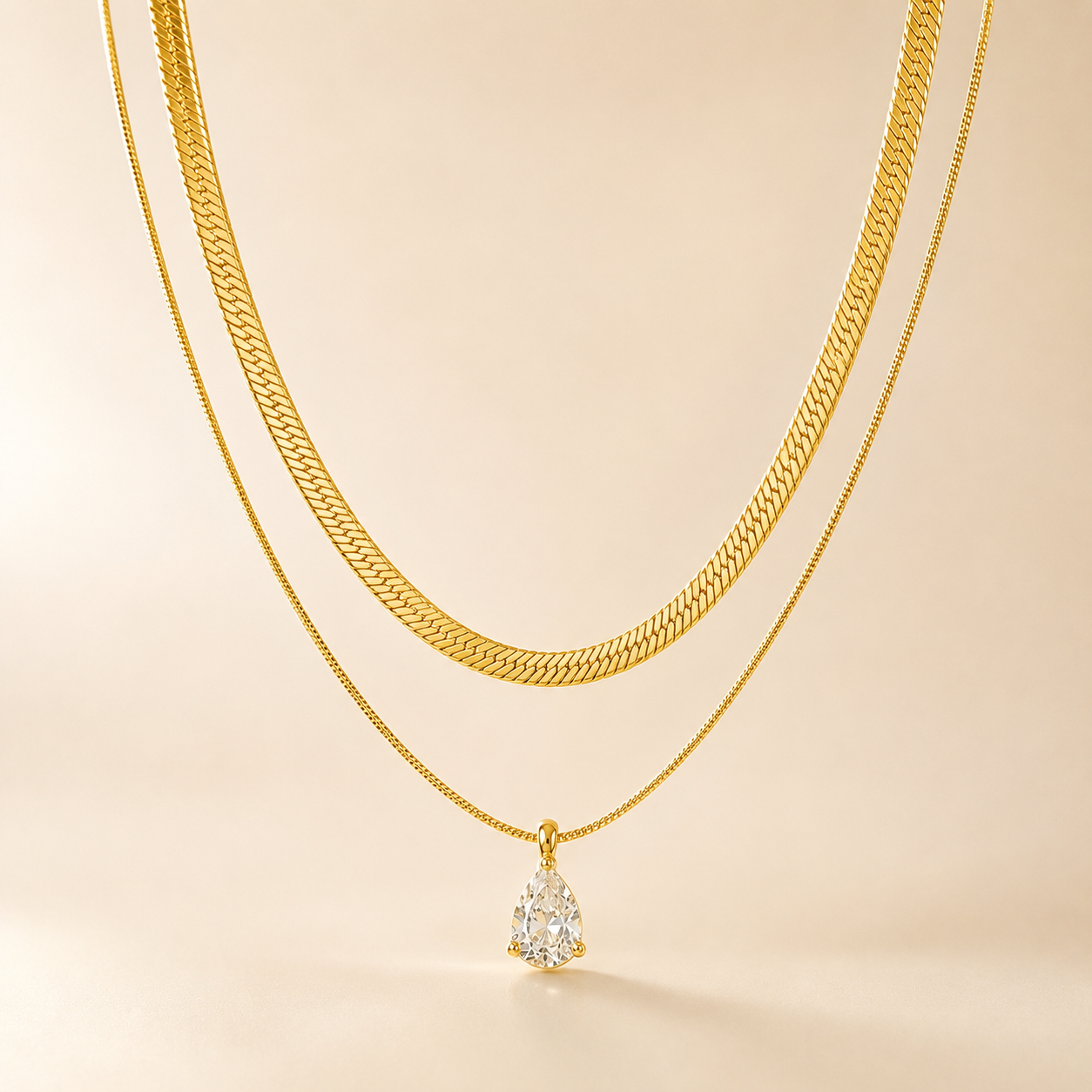 Gold-tone 316L layered necklace with a wide herringbone chain and a fine box chain holding a pear-cut clear stone pendant