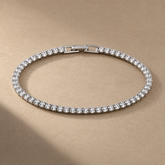 Silver-tone brass tennis bracelet with a row of clear round stones in claw settings and a box clasp, laid flat on a beige surface