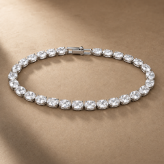 Silver-tone brass tennis bracelet with a row of oval clear stones in claw settings and a box clasp, laid flat on a beige surface