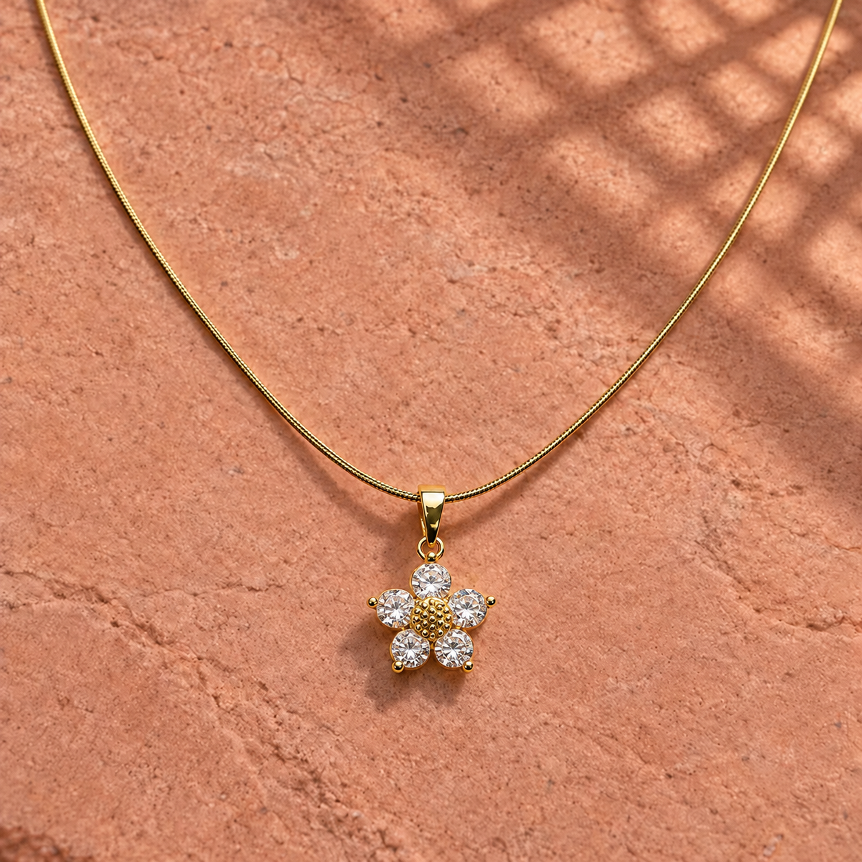 Gold-tone 316L stainless steel necklace with a snake chain and a flower pendant set with five clear stones, lying on a stone surface