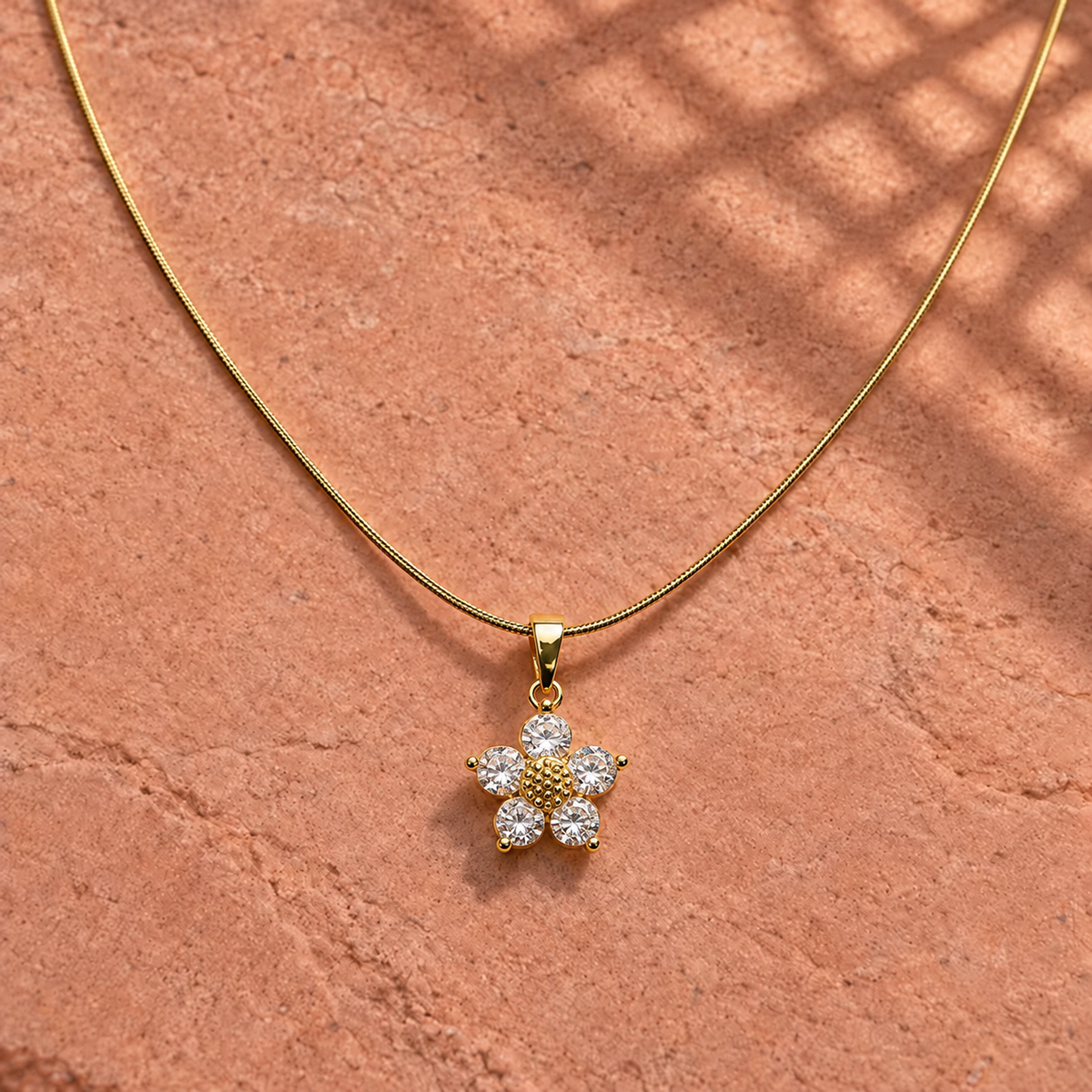 Gold-tone 316L stainless steel necklace with a snake chain and a flower pendant set with five clear stones, lying on a stone surface