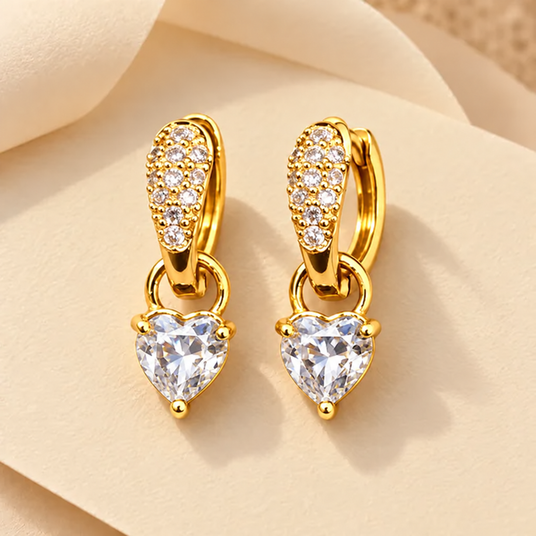Gold-tone 316L stainless steel huggie earrings with pavé crystal tops and a heart-cut crystal drop, a pair on a cream background