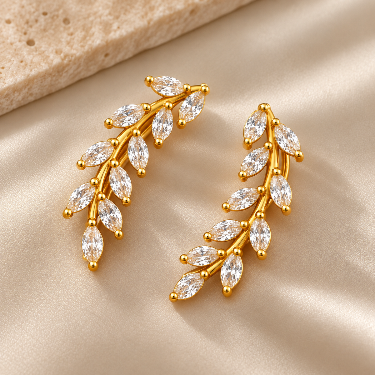 Gold-tone brass pair of leaf-spray earrings with marquise-cut clear stones along a curved stem, shown on a satin backdrop