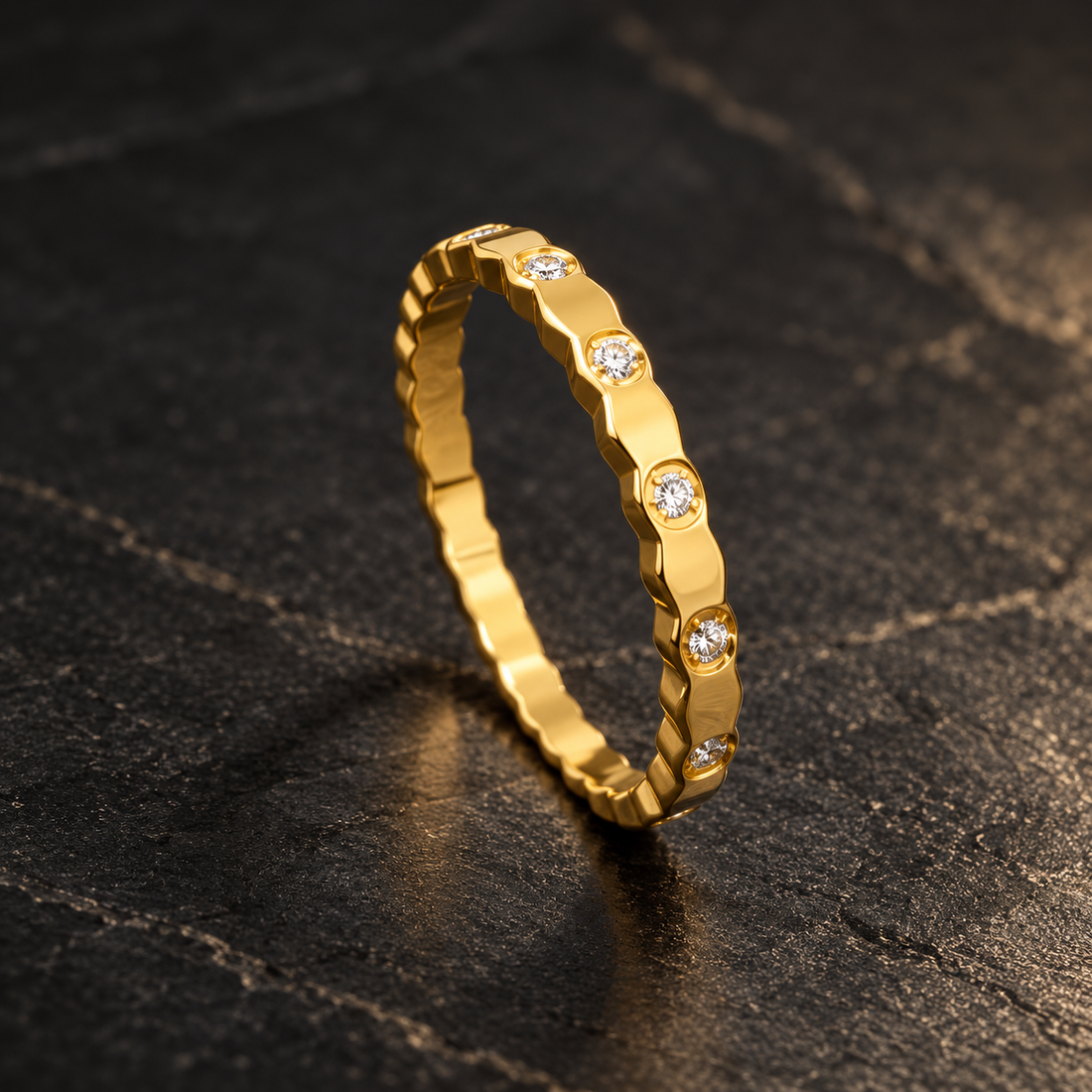 Gold-tone 316L stainless steel band ring with a faceted scalloped edge and clear crystals set at intervals, shown standing on a dark surface