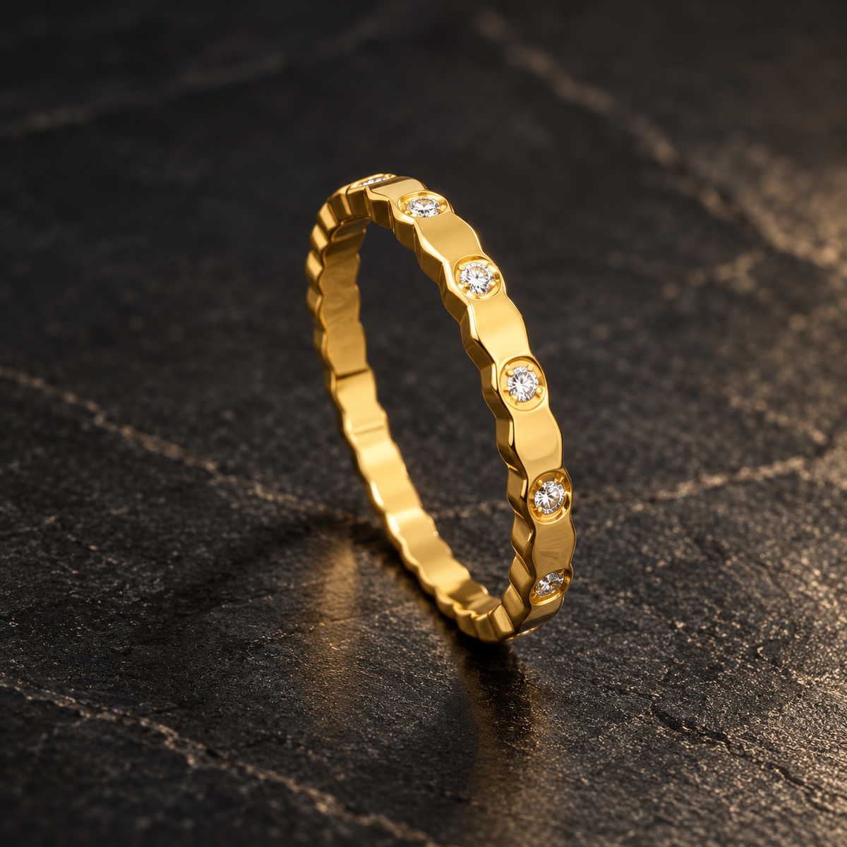 Gold-tone 316L stainless steel band ring with a faceted scalloped edge and clear crystals set at intervals, shown standing on a dark surface
