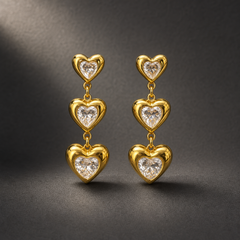 Two ear-worn earrings of one design in yellow-gold-tone metal with a polished finish. Each carries three solid-metal heart motifs of increasing size linked by jump rings, hinging at each junction so the sections swing independently; overall drop is roughly three times the width of the largest heart. Each heart is bezel-set with a clear heart-cut faceted stone, colourless to faintly warm, the stones sized to match their frames.