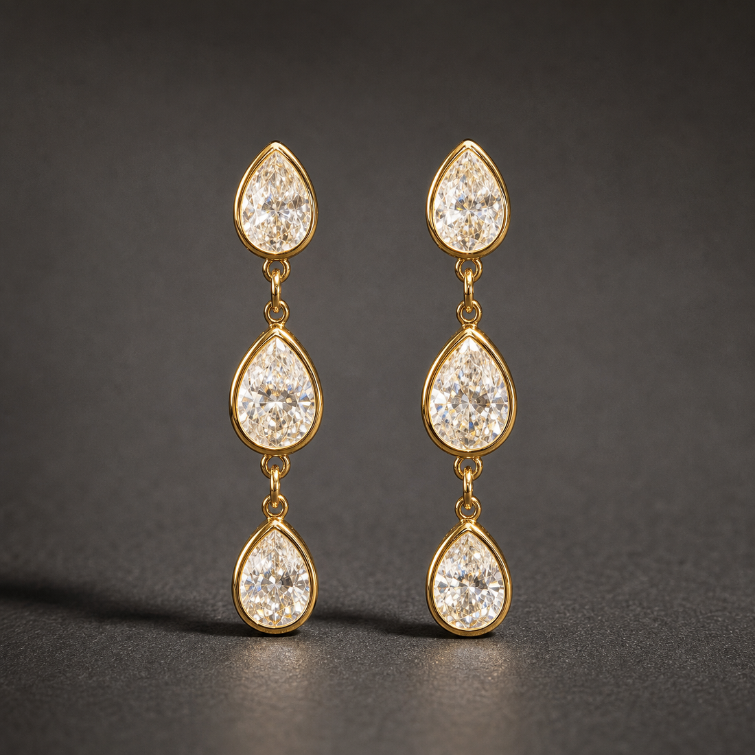 Two ear-worn earrings of one design in yellow-gold-tone metal with a polished finish. Each earring carries three pear-shaped clear faceted stones in matching gold-tone bezel-style mounts, linked vertically by small jump rings so the three sections articulate and swing independently. Stones decrease slightly in size downward, though the uppermost stone reads slightly smaller than the middle one. Overall drop is roughly four times the width of the widest stone.