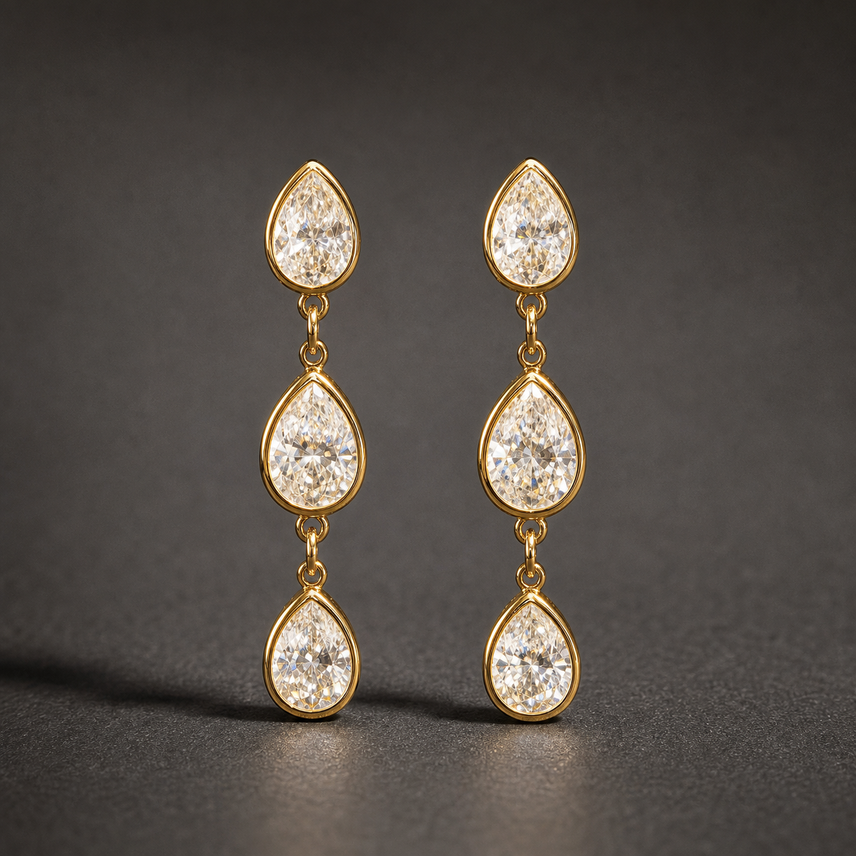 Two ear-worn earrings of one design in yellow-gold-tone metal with a polished finish. Each earring carries three pear-shaped clear faceted stones in matching gold-tone bezel-style mounts, linked vertically by small jump rings so the three sections articulate and swing independently. Stones decrease slightly in size downward, though the uppermost stone reads slightly smaller than the middle one. Overall drop is roughly four times the width of the widest stone.