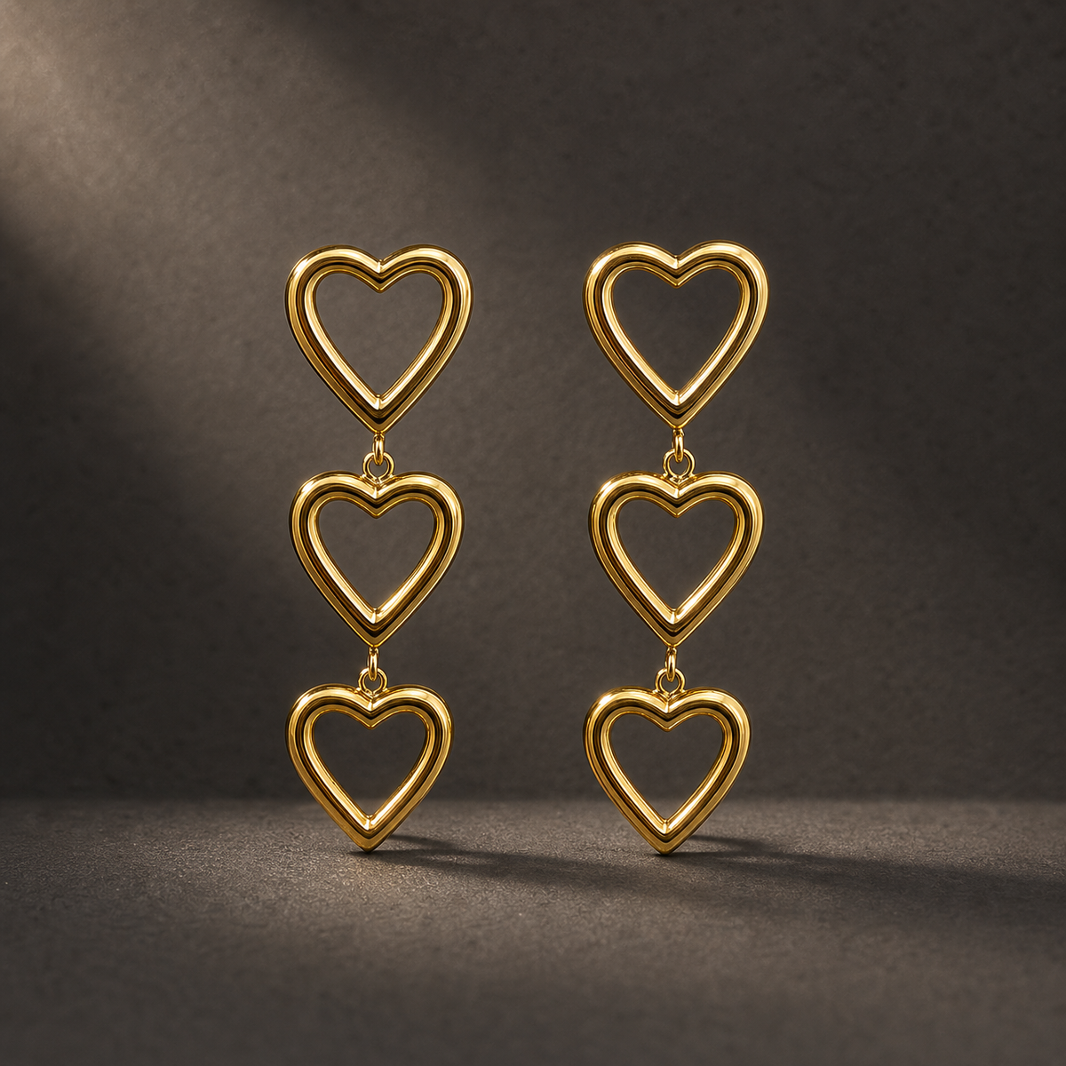Two ear-worn earrings of one design in yellow-gold-toned metal with a polished finish. Each is a three-tier drop of open-frame solid-metal heart motifs, roughly equal in size, the top heart carrying the fitting and each lower heart suspended from the one above by small jump-ring links, giving three articulated sections; overall drop is approximately three times the width of the widest heart.