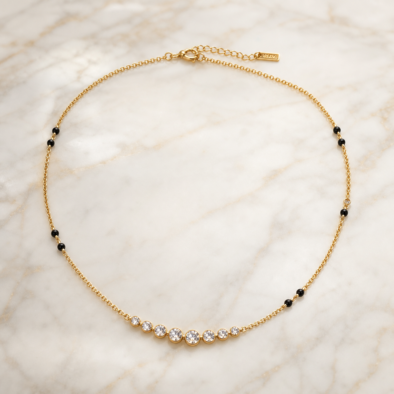 One single-strand necklace in yellow-gold-tone metal with a polished finish, built on a fine-gauge cable chain. Along the strand from the clasp end: the clasp assembly comprises a spring-ring or lobster clasp with a short extender chain terminating in one rectangular stamped metal tag; moving along the chain there are spaced stations of small round black beads, mounted directly on the chain, appearing in adjacent pairs with uneven gaps between pairs — one pair, then a long plain run, then another pair,