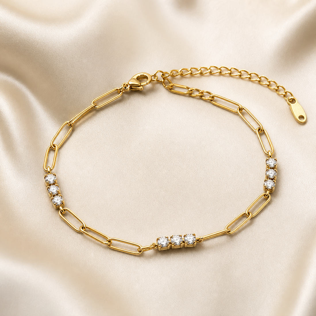 One flexible bracelet in yellow-gold-toned metal with a polished finish, built as a single strand of elongated paperclip-style oval links of medium gauge. From the clasp end: a lobster clasp terminates the far end; moving along the strand, a run of plain paperclip links leads to the first station of three round clear faceted stones, each in a four-prong setting, closely spaced in a short straight line; another plain paperclip section follows, then a second identical three-stone station; more paperclip