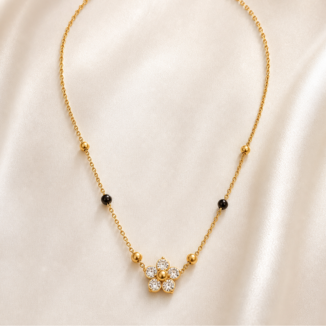 A single gold-tone stainless steel necklace with a polished finish, built on one fine-gauge cable chain strand. From the clasp end: a lobster clasp with a short oval-link extender chain terminating in a small rectangular bar tag; the plain chain continues to the decorative section, where fixed bead stations appear in sequence on each side — first a small round polished gold bead, then a small round black bead, then another small round gold bead — giving four gold beads and two black beads in total, all