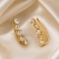 A matched pair of ear climber earrings, two items, in polished yellow-gold-coloured metal. Each earring comprises a slender curved wire frame forming an elongated open hook with a long plain tail that follows the ear, fronted by a graduated line of prong-set clear faceted stones fixed directly to the frame.