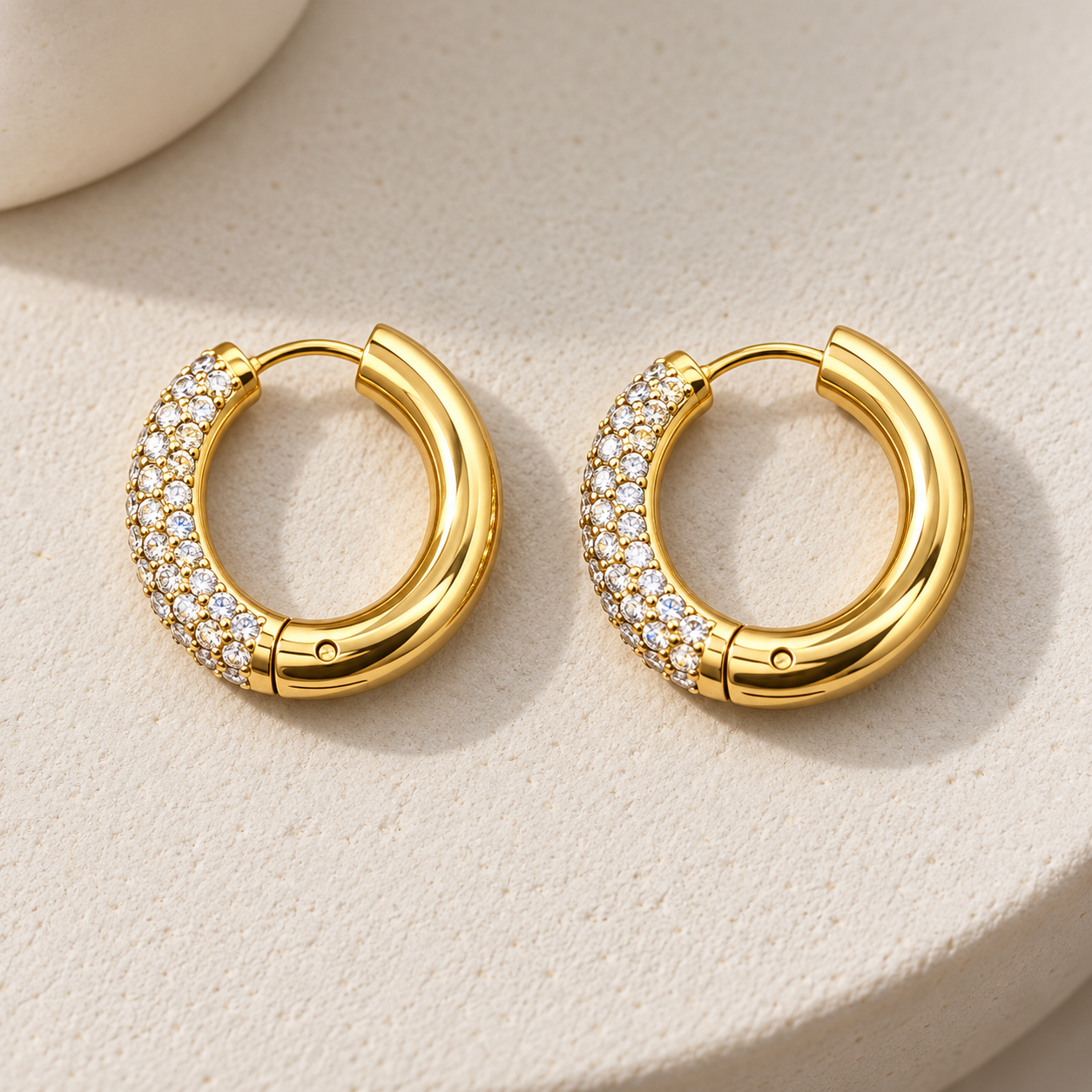 Gold-tone 316L stainless steel pair of hinged hoop earrings with clear stones set across the front third of each hoop, shown on a cream surface