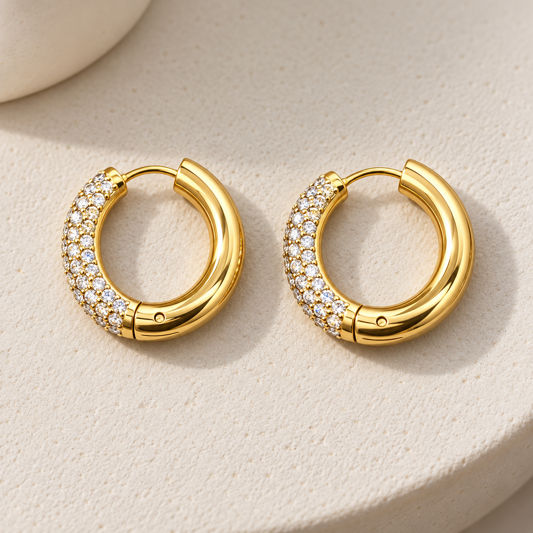 Gold-tone 316L stainless steel pair of hinged hoop earrings with clear stones set across the front third of each hoop, shown on a cream surface