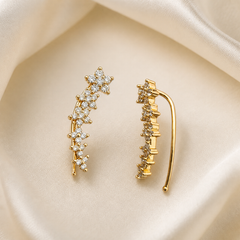 A matched pair of ear-climber earrings in yellow gold with a polished finish. Each earring consists of a narrow curved metal spine following the ear's contour, terminating at the lower end in a thin open shepherd's-hook ear wire with a small ball tip. Along the outer face of the spine, from the top down toward the hook, runs an irregular trail of small clear round faceted stones of graduated and mixed sizes, each individually held in raised prong settings; the stones form clustered groupings rather than an