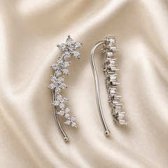 A matched pair of ear-climber earrings, shown from both the decorative face (one earring) and the reverse (the other). The metal is silver-toned with a polished finish. Each earring is a single rigid curved stem of slim wire that hooks over the ear, carrying a graduated line of floral clusters along its outer edge; the reverse shows a series of open box-style prong settings in a stepped row.