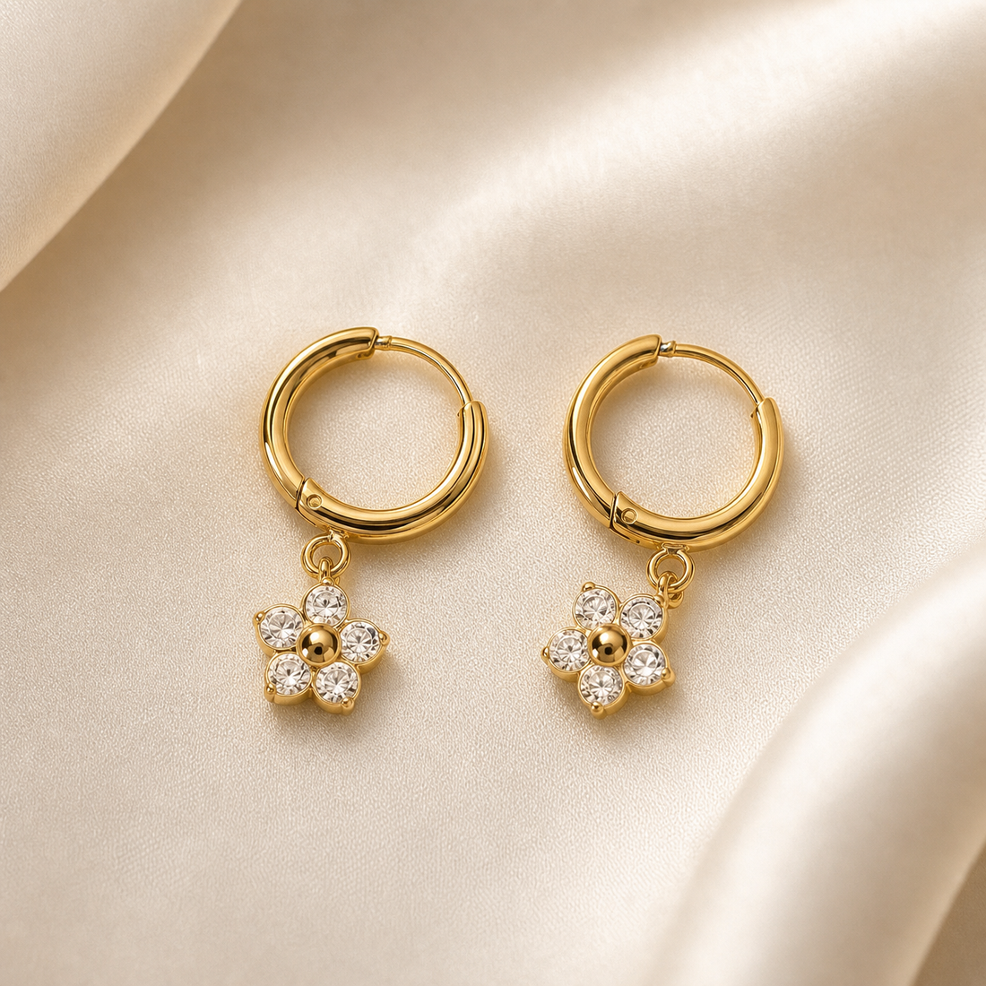 A matched pair of two drop earrings in yellow-gold-toned metal with a polished finish. Each earring consists of a small round huggie-style hoop with a hinged click closure, from which a single flower charm dangles on a short twisted jump-ring connector. Each flower charm is a solid-metal five-petal motif in the same gold tone, with each rounded petal set with a clear round faceted stone in a flush or bezel-style mounting, and a small polished gold dome or bead forming the raised centre; the stones are