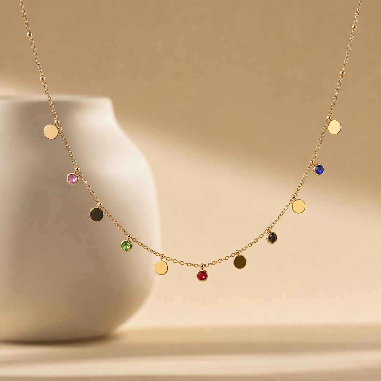 One station necklace in gold-tone metal with a polished finish, built as a single fine cable chain of small oval links. From the clasp end along the strand: a small gold bead, then alternating dangling charms — a gold disc, a small pink round stone in a bezel cup, a dark disc, a green round stone, a gold disc, a central red round stone, a dark disc, a dark stone, a gold disc, a blue round stone, and a final gold disc toward the far end. Charms drop from jump-ring or short-stem mountings, spacing irregular.