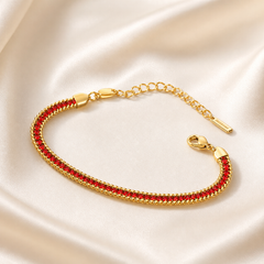 A single flexible bracelet in yellow-gold-toned metal with a polished finish. The main body is a flat, tightly woven herringbone-style chain of fine gauge, bordered on both long edges by a slightly raised twisted rope trim. Set continuously along the centre of the band is one unbroken row of small round faceted red stones, each held in an individual metal bezel or channel setting, the settings matching the gold tone of the chain; the row runs the full decorative length between two smooth, shield-shaped