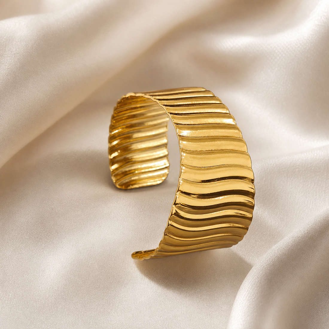 A single wide cuff bracelet in polished yellow-gold-toned metal. The band is an open, rigid cuff of broad, slightly tapering width, its outer surface formed as continuous horizontal ribbing — a repeating series of raised, softly undulating ridges and recessed channels running across the full width of the band, giving a corrugated, wave-like relief. The finish is bright and mirror-polished on the ridge crests, with the grooves reading darker.