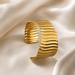 A single wide cuff bracelet in polished yellow-gold-toned metal. The band is an open, rigid cuff of broad, slightly tapering width, its outer surface formed as continuous horizontal ribbing — a repeating series of raised, softly undulating ridges and recessed channels running across the full width of the band, giving a corrugated, wave-like relief. The finish is bright and mirror-polished on the ridge crests, with the grooves reading darker.