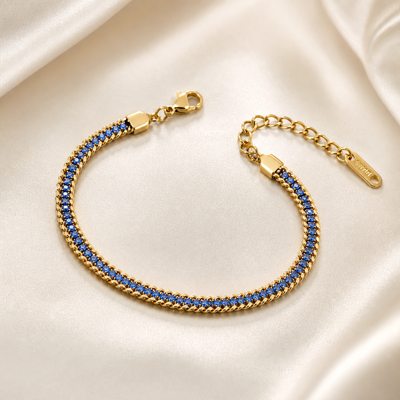 A single flexible bracelet in yellow gold tone with a polished finish. The central body is a continuous single strand of round faceted blue stones set in a straight line, each stone of uniform small size, mounted within a narrow polished gold channel flanked on both long edges by fine curb-chain trim of small gold links, so the strand reads as a stone line bordered by chain.