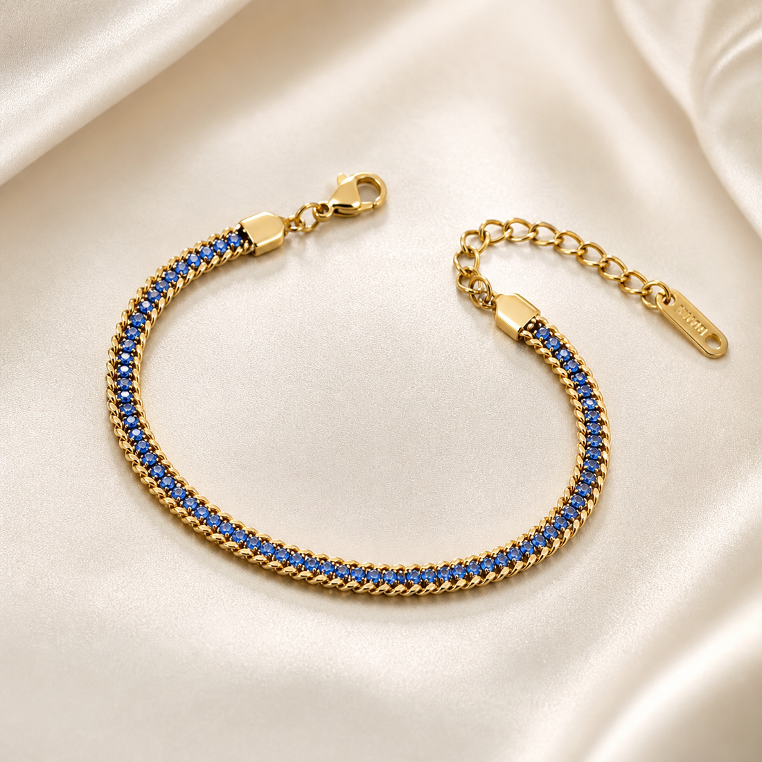 A single flexible bracelet in yellow gold tone with a polished finish. The central body is a continuous single strand of round faceted blue stones set in a straight line, each stone of uniform small size, mounted within a narrow polished gold channel flanked on both long edges by fine curb-chain trim of small gold links, so the strand reads as a stone line bordered by chain.