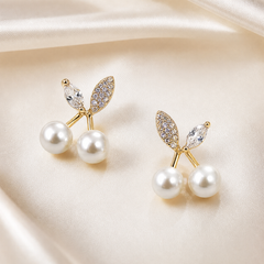 A matched pair of stud earrings, two items total. Yellow-gold-tone metal with a polished finish. Each earring is built as a small cherry cluster on a single stud post: two short gold stems branch from a shared base, each stem terminating in a round white faux-pearl bead of medium size, mounted as a peg-set stud bead; from the same base extend two leaf motifs.
