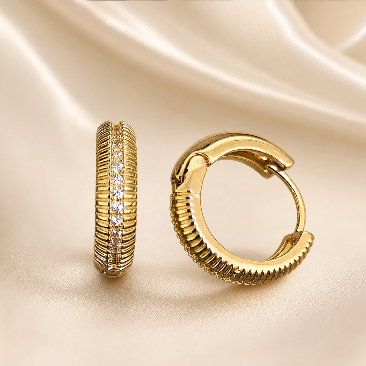 A matched pair of small hinged huggie hoop earrings, two items total. Yellow-gold-toned metal with a polished finish. Each earring is a compact, thick hoop of rounded cross-section; the outer face carries fine horizontal ribbing (closely spaced reeded grooves) running around the curve, interrupted by a single straight central channel set with a continuous line of small, round, clear faceted stones held in a narrow pavé-style metal setting. The inner portion of each hoop is smooth polished metal.