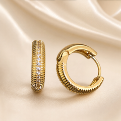 A matched pair of small hinged huggie hoop earrings, two items total. Yellow-gold-toned metal with a polished finish. Each earring is a compact, thick hoop of rounded cross-section; the outer face carries fine horizontal ribbing (closely spaced reeded grooves) running around the curve, interrupted by a single straight central channel set with a continuous line of small, round, clear faceted stones held in a narrow pavé-style metal setting. The inner portion of each hoop is smooth polished metal.