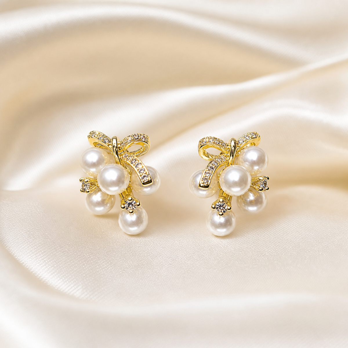 A matched pair of stud earrings in gold-tone metal with a bright polished finish. Each earring is built as a ribbon-bow motif whose two loops and two short tails are set with small clear pavé stones, fixed above a compact dangling cluster of round white imitation pearls of graduated sizes, with one noticeably larger central pearl and smaller pearls grouped around it; tiny clear stone accents sit at the cluster edges.
