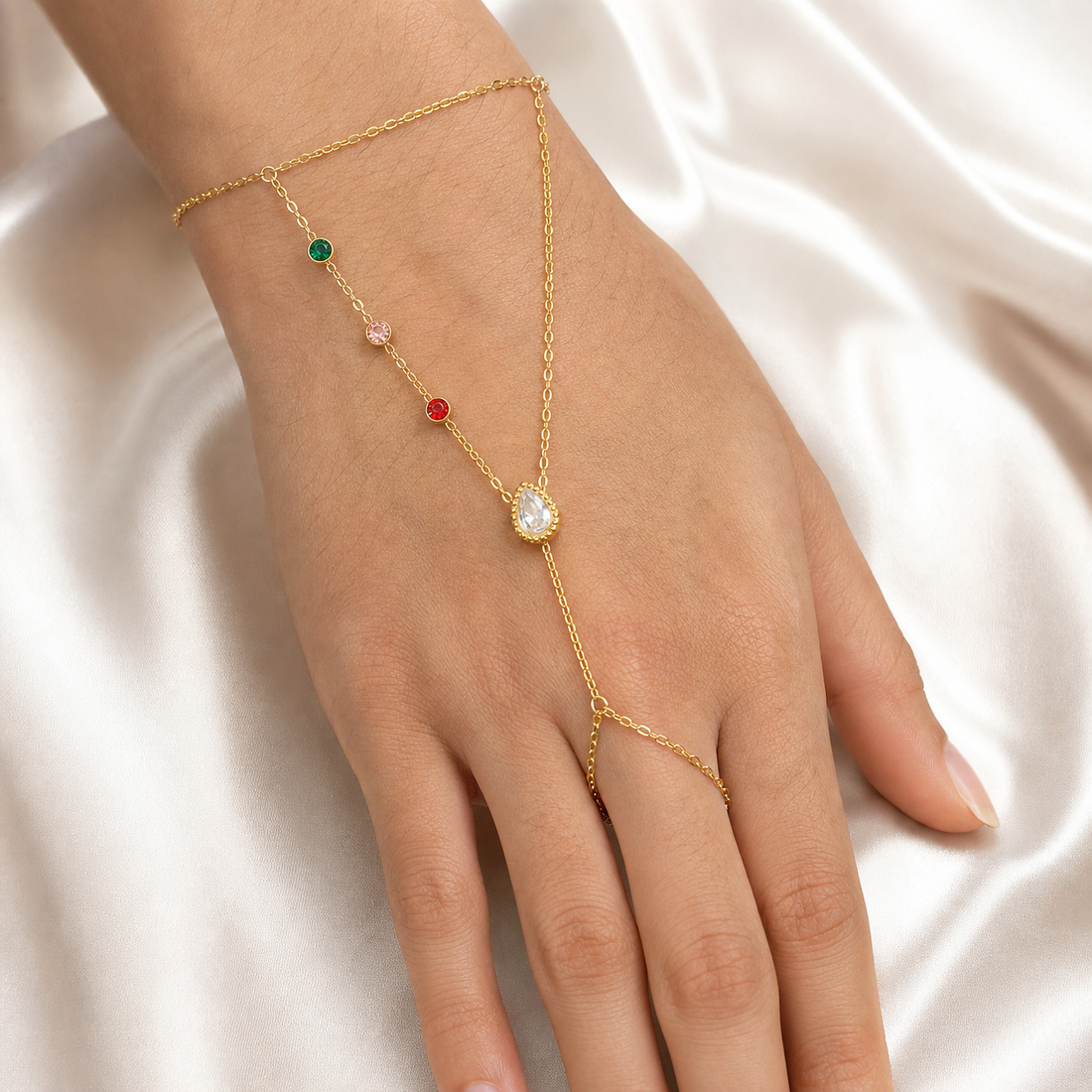 One gold-tone hand-chain piece with a polished yellow-metal finish. A single fine cable-chain wrist bracelet forms the fixed wrist section; its clasp, extender and any terminal tag are not clearly visible and are treated as obscured. Two fine-gauge cable-chain branches attach to the wrist section and converge at one central junction: the first branch carries, in sequence from the wrist, three small round bezel-set stones coloured green, light pink and red, each individually separated by short chain runs;