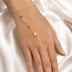 One gold-tone hand-chain piece with a polished yellow-metal finish. A single fine cable-chain wrist bracelet forms the fixed wrist section; its clasp, extender and any terminal tag are not clearly visible and are treated as obscured. Two fine-gauge cable-chain branches attach to the wrist section and converge at one central junction: the first branch carries, in sequence from the wrist, three small round bezel-set stones coloured green, light pink and red, each individually separated by short chain runs;