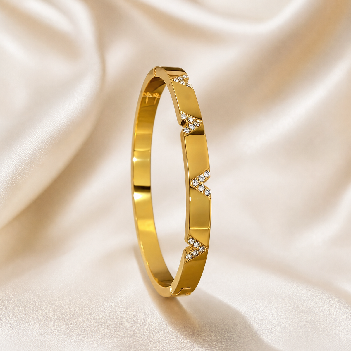 A single rigid bangle bracelet in yellow-gold-toned metal with a high-polish mirror finish. The band is a continuous closed oval of medium width with flat outer faces and squared edges. Around the outer face run repeated V-shaped chevron notches cut into the band, each chevron recess filled with small round clear faceted stones in pavé-style settings; the stone-filled chevrons alternate with plain polished gold segments of the band.