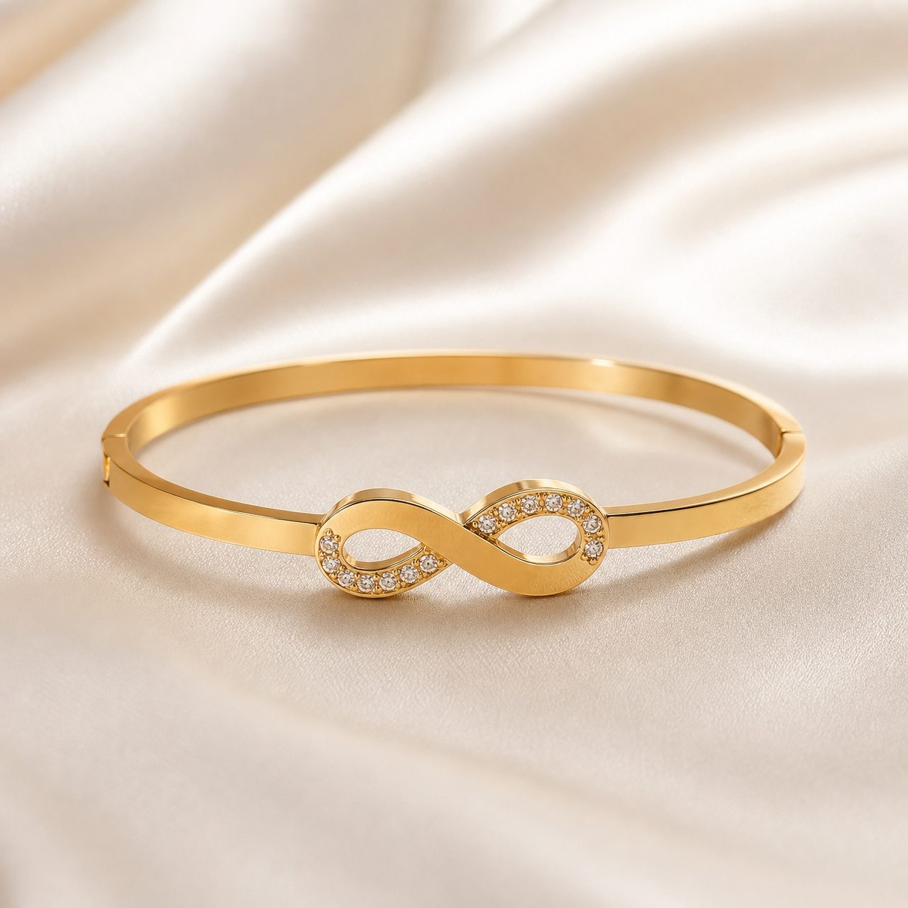 One rigid bangle bracelet in warm yellow-gold-toned metal with a polished, reflective finish. The piece is a narrow, flat-sided band of even gauge forming a closed oval, with a visible hinge seam on one side of the band and a small notched closure seam on the opposite side. The sole decorative component is a single solid-metal infinity (figure-eight) motif mounted centrally as a fixed top feature, joined directly to the band at both of its outer loops.