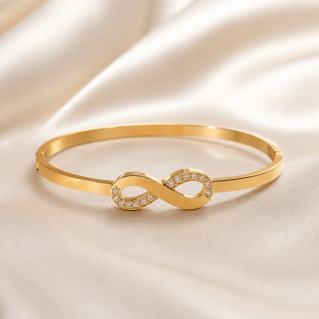 One rigid bangle bracelet in warm yellow-gold-toned metal with a polished, reflective finish. The piece is a narrow, flat-sided band of even gauge forming a closed oval, with a visible hinge seam on one side of the band and a small notched closure seam on the opposite side. The sole decorative component is a single solid-metal infinity (figure-eight) motif mounted centrally as a fixed top feature, joined directly to the band at both of its outer loops.