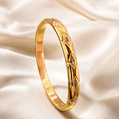 A single rigid bangle (kada) in yellow gold with a high-polish finish and a warm rose-gold reflection on its inner band. The band is a continuous closed oval of medium gauge with a smooth, plain interior and a decorated outer face. The outer surface carries a repeating engraved relief of pointed marquise-shaped leaf or eye motifs linked by raised polished ribs; each motif is a solid-metal frame flush-set with tiny round stones.