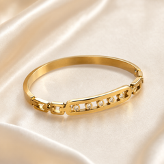 A single rigid bangle in yellow-gold-toned metal with a high-polish finish. The band is a solid, smooth, slightly flat-profiled hoop of even width around most of its circumference, interrupted by one decorative section and a hinged box-style clasp with a safety catch.
