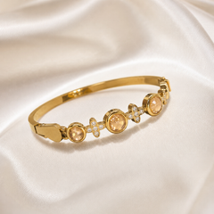 One rigid bracelet in polished yellow gold with a high-gloss finish, formed as a smooth, plain narrow band around most of its circumference with a decorated front section. The decorative front carries, in sequence, three round champagne-peach faceted stones in smooth raised bezel settings, alternating with two small four-petal flower motifs, each petal set with tiny clear faceted stones in a pavé-style mounting, the flowers set between and flanking the bezels.