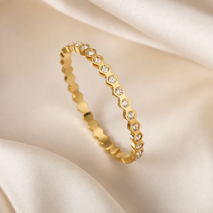 A single rigid bangle, one item, in yellow-gold-toned metal with a high-polish finish. The band is of medium, even gauge with a continuous scalloped outer edge formed by a repeating series of rounded bezels, each holding one clear round faceted stone set flush in a bezel mounting; the stones are uniform in size and colourless, spaced evenly and continuously around the circumference so that metal scallops separate each stone from the next. The inner edge of the band is smooth and plain.