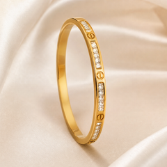 A single rigid bangle of yellow-gold-coloured polished metal, circular and closed with no visible clasp, hinge or opening mechanism resolved. The band is of narrow, even gauge with a smooth domed profile. Around its outer face, solid-metal screw-head motifs — small circular discs each bearing a recessed horizontal slot — alternate with short channel-set rows of clear square-cut faceted stones set flush into the band.
